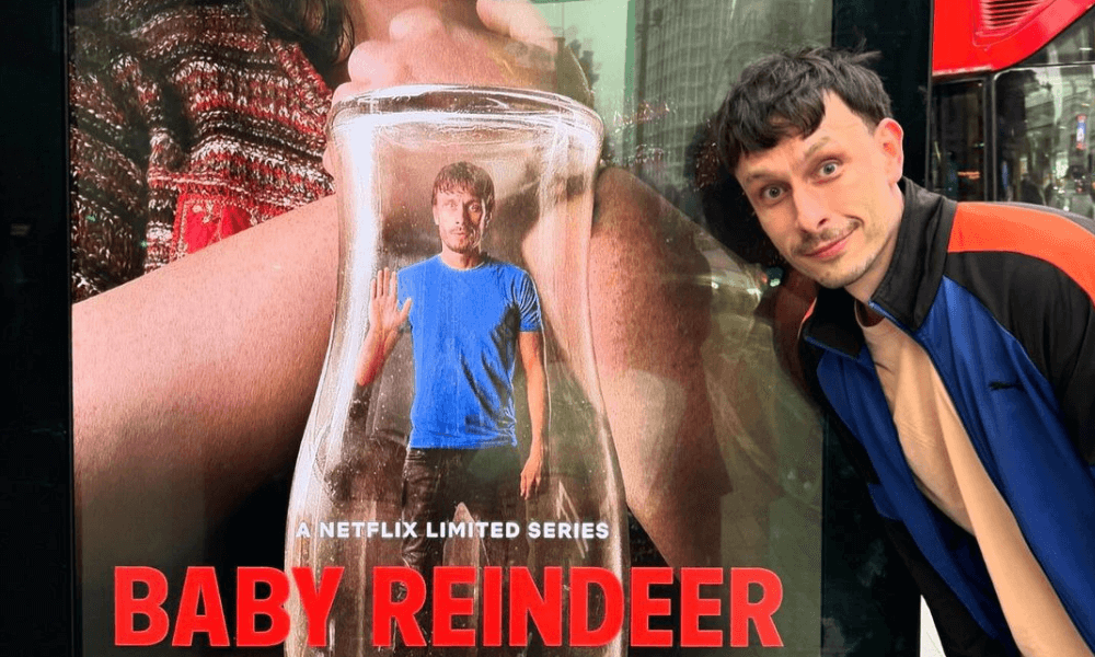 A man poses beside a bus stop advertisement for the netflix series "baby reindeer," which shows another man inside a large jar.