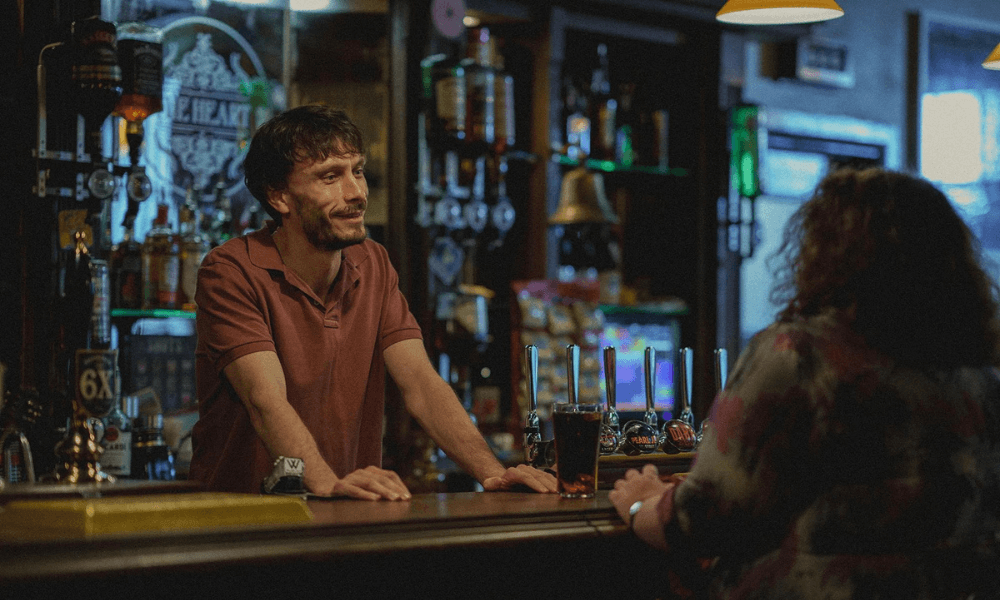 A bartender converses with a female customer at a dimly lit bar, with drinks and assorted bottles in the background.