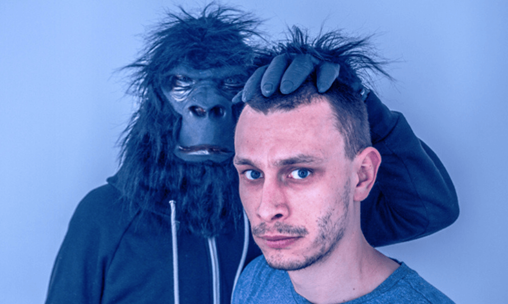 A man standing next to another person wearing a gorilla mask, who is placing a hand on the man's head, both against a light blue background.