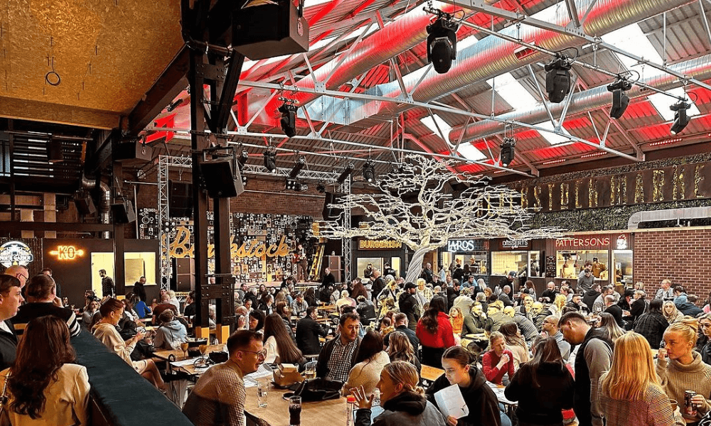 Crowded food market hall with patrons dining under a glass ceiling and decorative lighting, with food stalls and a tree sculpture in the background.