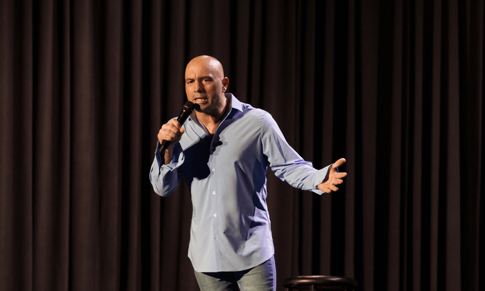 A bald man in a blue shirt performing stand-up comedy on stage with a microphone, gesturing with his right hand, in front of a dark curtain.