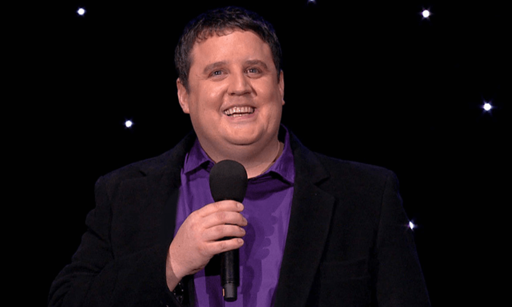 Man with microphone smiling on stage with starry background. he is wearing a purple shirt and suit.