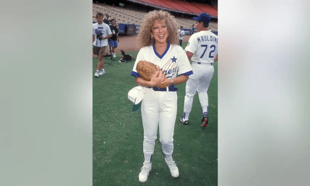 A woman with curly blonde hair, wearing a dodgers baseball uniform and holding a mitt, stands smiling on a baseball field.