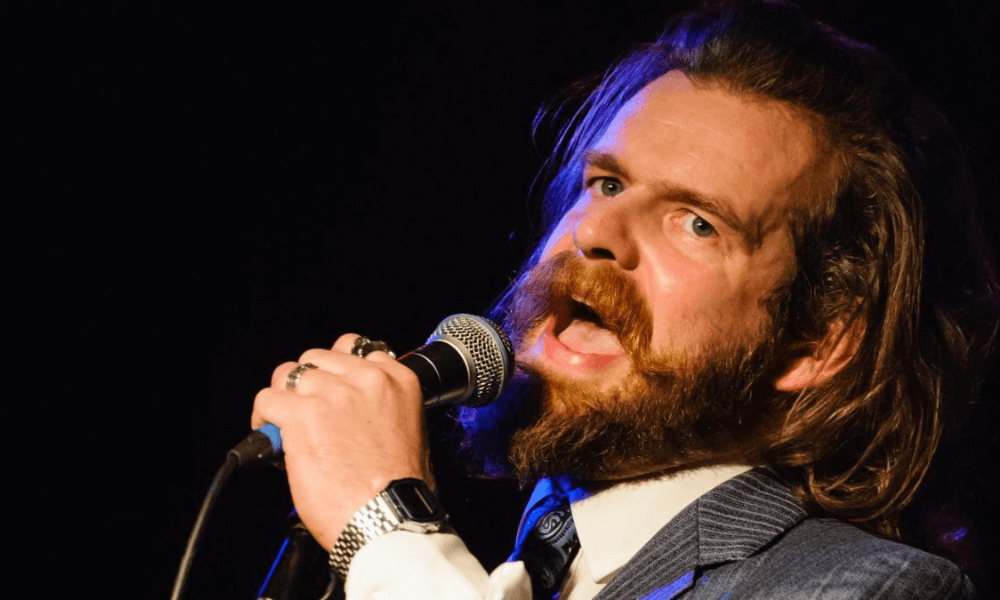 A bearded man singing into a microphone on stage, wearing a suit and tie, with intense expression and long hair.