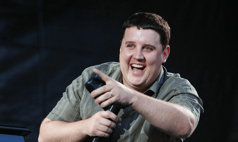 A jovial man holding a microphone, laughing while speaking onstage, dressed in a plaid shirt.
