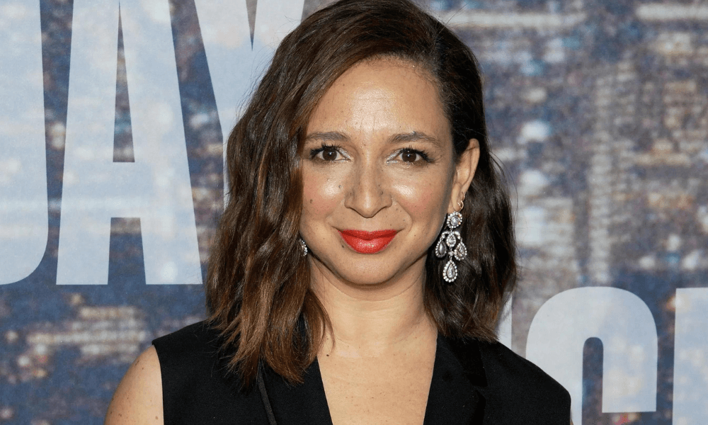 Maya rudolph smiling at a premiere event, wearing a black blazer and dangling earrings, with a movie backdrop behind her.