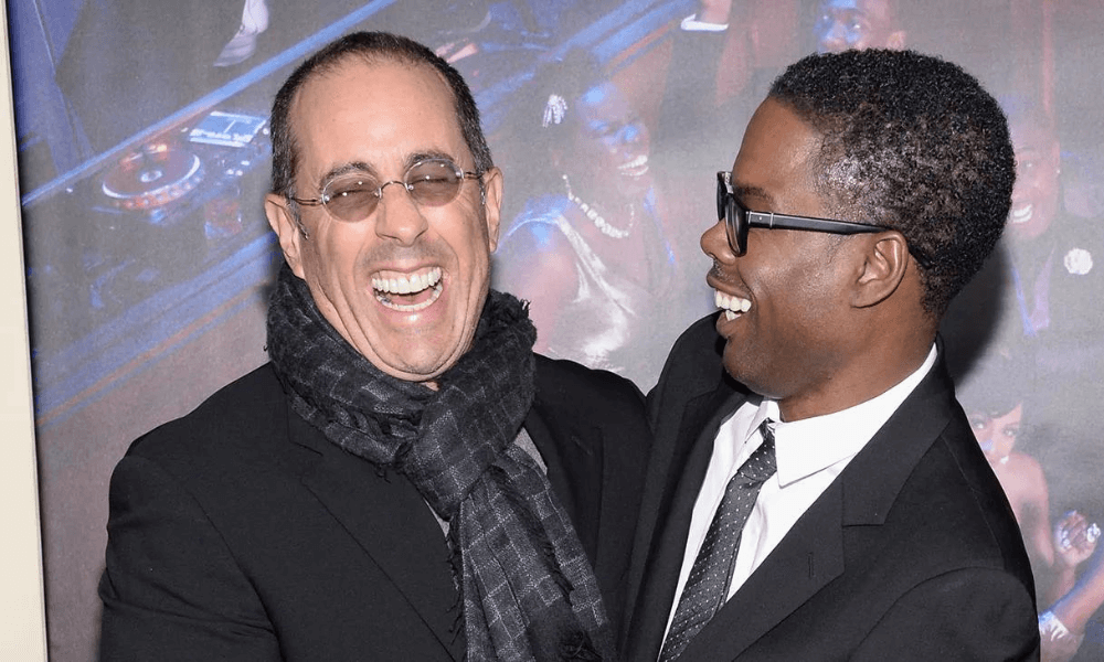 Two men laughing together at a movie premiere, one wearing a black scarf and coat, the other in a classic suit with glasses.