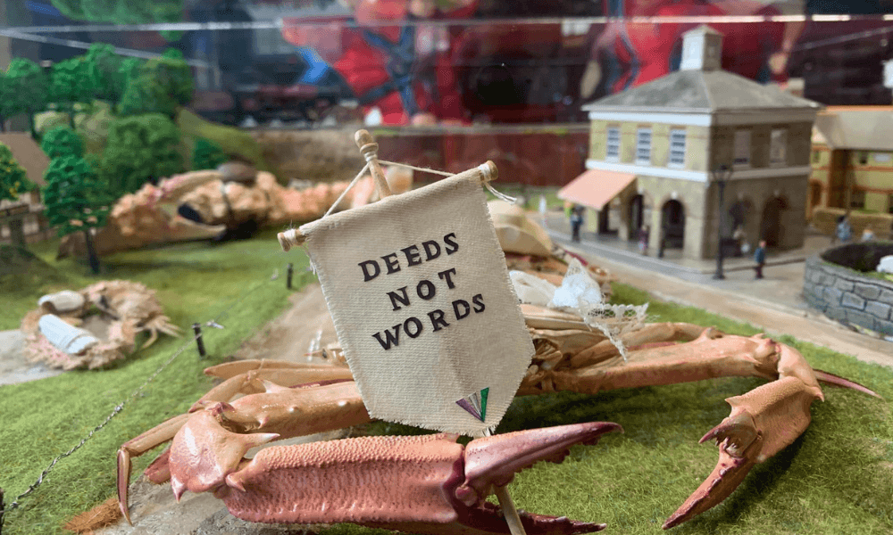 Miniature scene of crabs holding a banner reading "deeds not words" in a display case with a small model town background.