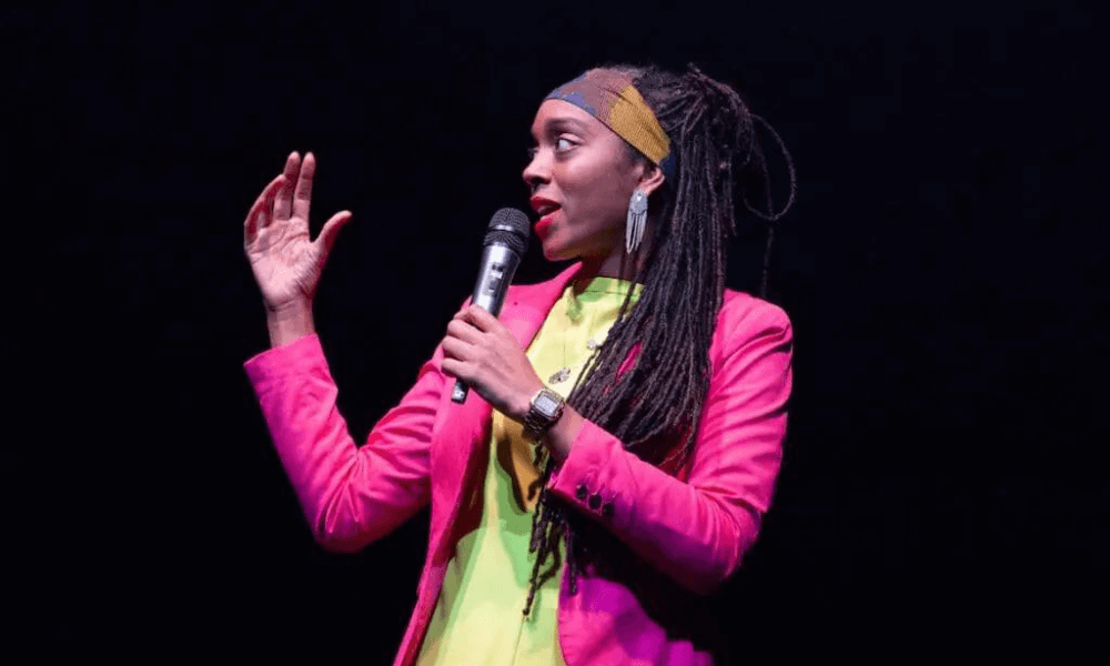 Woman presenting on stage, wearing a bright pink jacket and yellow shirt, holding a microphone, gesturing with her right hand.