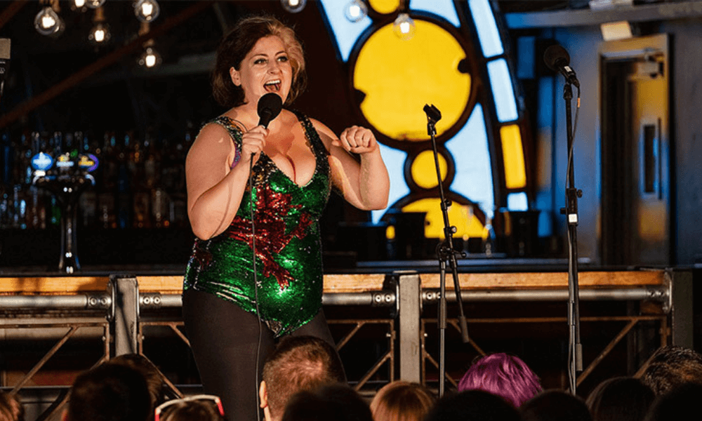 A woman in a sequined top performs stand-up comedy on stage in a bar, with an audience in the foreground and drinks in the background.