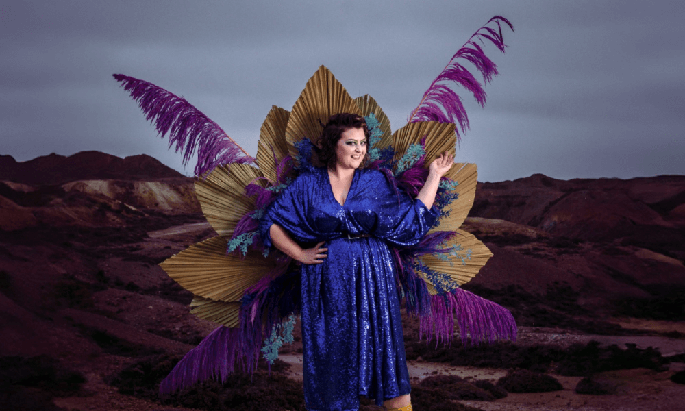 A woman in a sparkling blue dress and large, colorful feathered wings poses dramatically against a muted desert landscape.