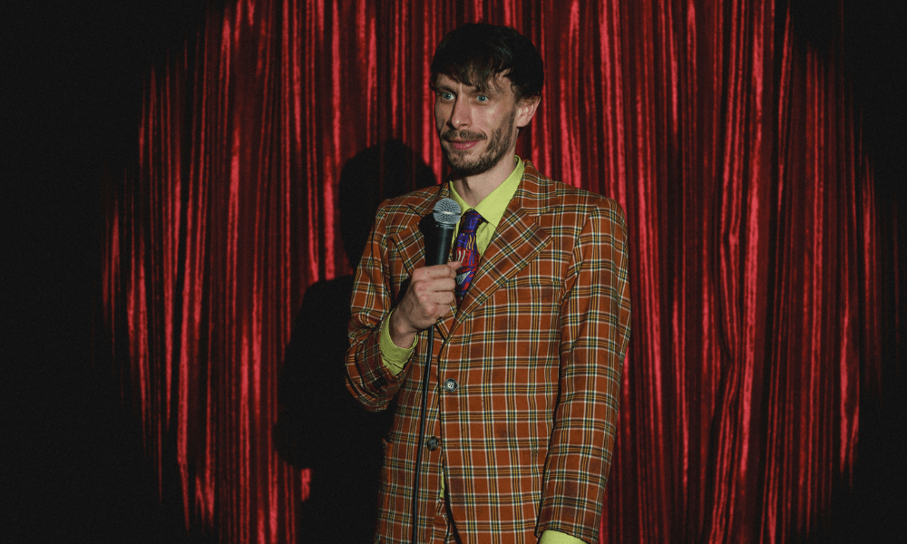 A man in a plaid suit stands with a microphone against a red curtain background, smiling slightly.