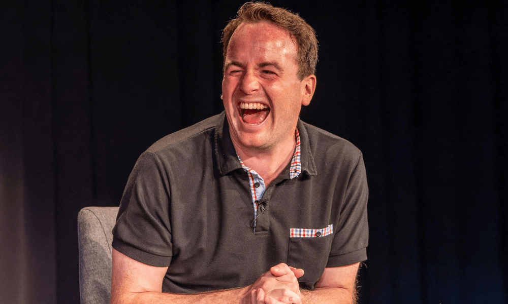 Man laughing heartily while seated on stage, wearing a dark polo shirt with a light-colored handkerchief in the pocket.