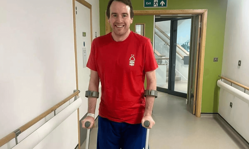 A man in a red t-shirt and black shorts using crutches stands smiling in a hospital corridor next to a green exit sign.