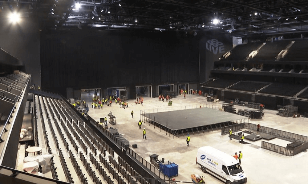 Workers setting up a large stage in an indoor arena with rows of seating and lighting equipment overhead.