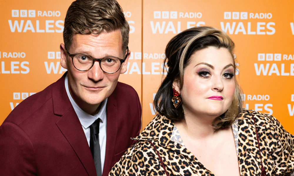 A man in a maroon suit and a woman in a leopard print top pose in front of a bbc radio wales backdrop.