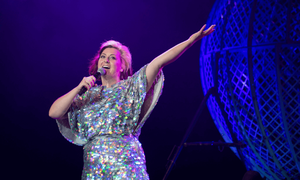 A woman with colorful hair, wearing a sparkling dress, performs on stage, gesturing with one arm raised, with a large blue spherical cage in the background.