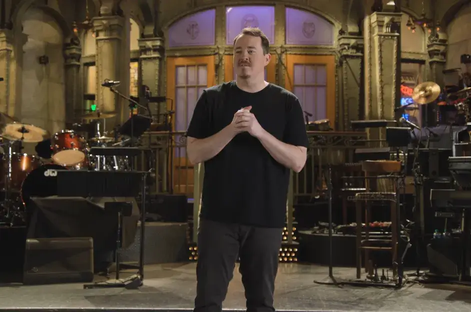 A man in a black t-shirt stands in front of a stage set with musical instruments, his hands clasped together.
