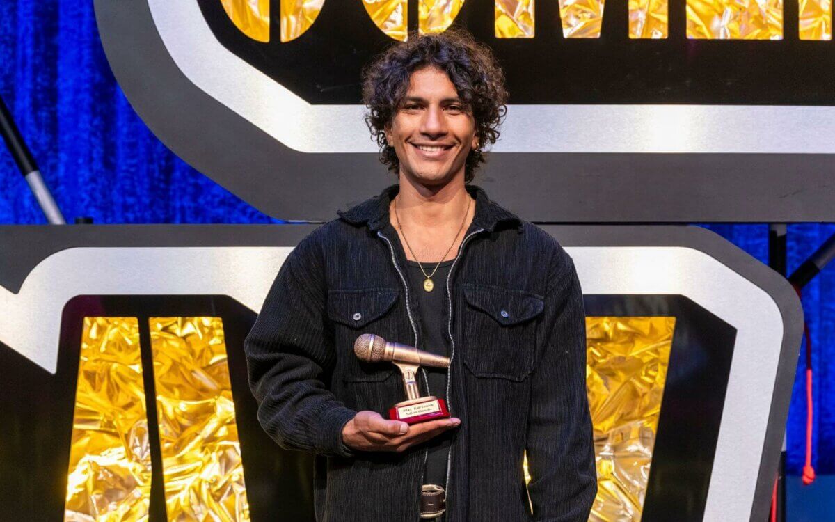 A young man with curly hair, smiling, holding a trophy on a stage with a gold and blue backdrop.