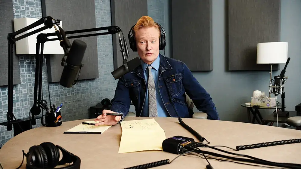A man with red hair in a denim jacket sits at a podcasting desk with microphone, speaking and writing notes.