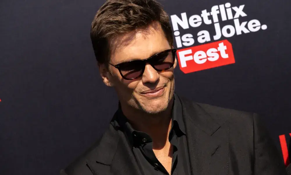 Man in sunglasses smiling at a "netflix is a joke fest" red carpet event. he wears a black suit and has short hair.
