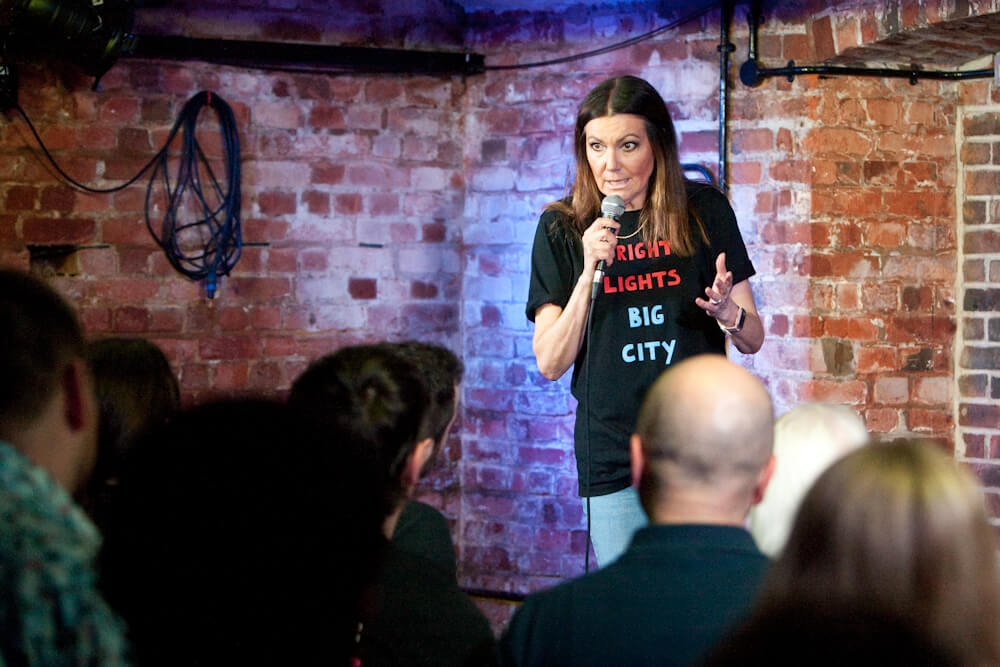 A woman speaking into a microphone at a stand-up comedy event, wearing a "bright lights big city" t-shirt, with an audience in the foreground and a brick wall background.