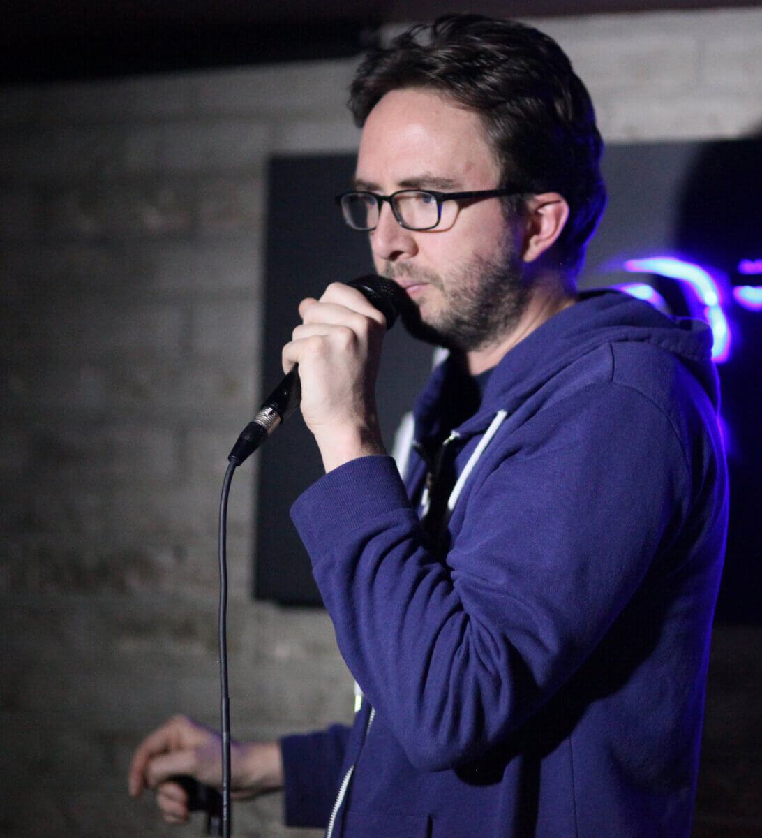 A man in glasses and a blue hoodie performing at a microphone with dim lighting and neon lights in the background.