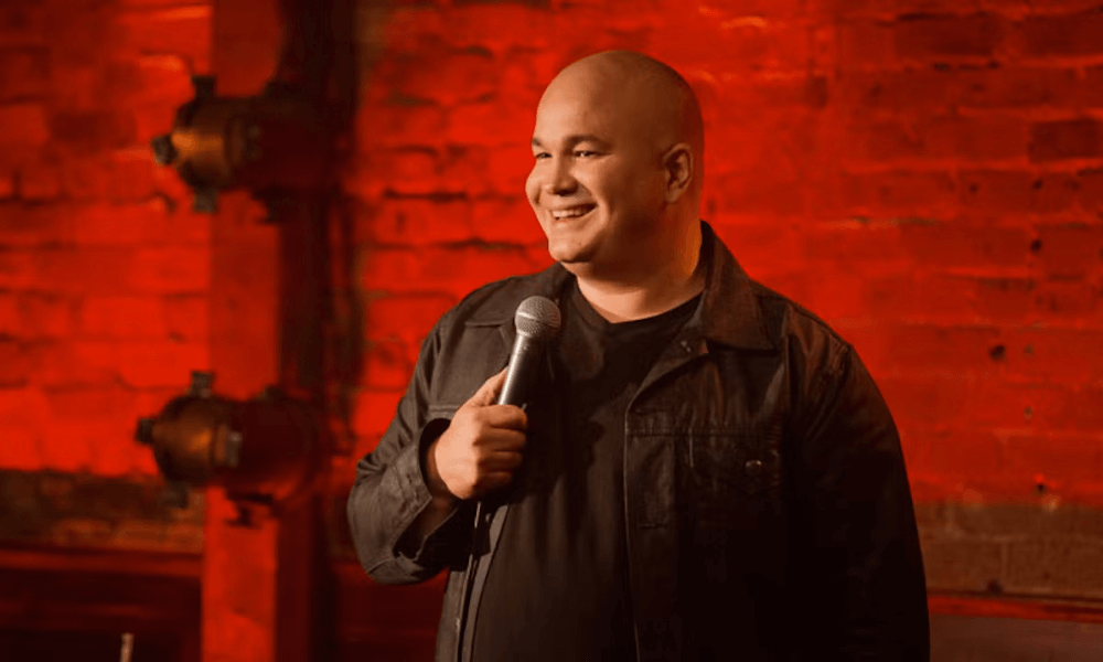 A bald man with a microphone smiling while performing stand-up comedy in front of a brick wall.