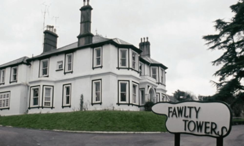 A large white traditional building with multiple chimneys, surrounded by trees, and a sign in the foreground reading "fawlty tower.