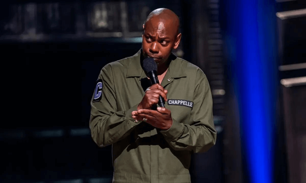 Comedian on stage performing into a microphone while wearing an olive green jacket with his last name, "chappelle", embroidered on it.