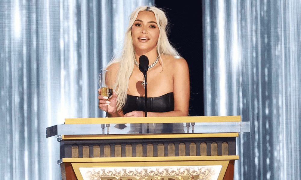 Woman with long blonde hair, wearing a black strapless top, speaking at a podium with a microphone, holding an award.
.