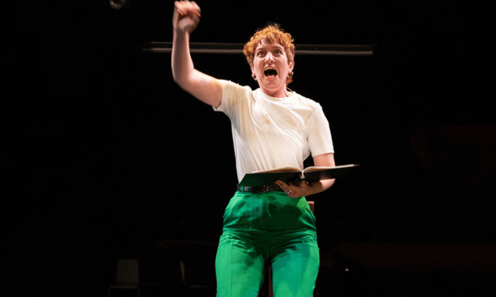 A woman on stage, expressive and holding a book, wears a white t-shirt and green pants as she raises her left arm.