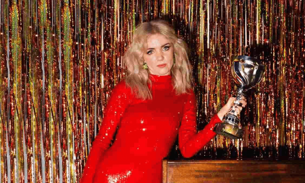 A woman in a red dress holding a trophy, standing in front of a shimmering multicolored backdrop.