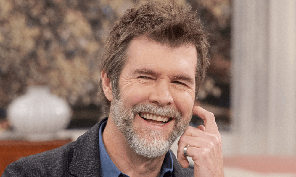 A middle-aged man with salt-and-pepper beard and hair smiling warmly, sitting with his finger on his cheek. he wears a grey blazer over a blue shirt.