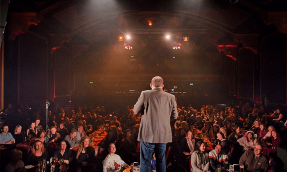 A man stands on stage speaking to a crowded theater audience under warm stage lighting.