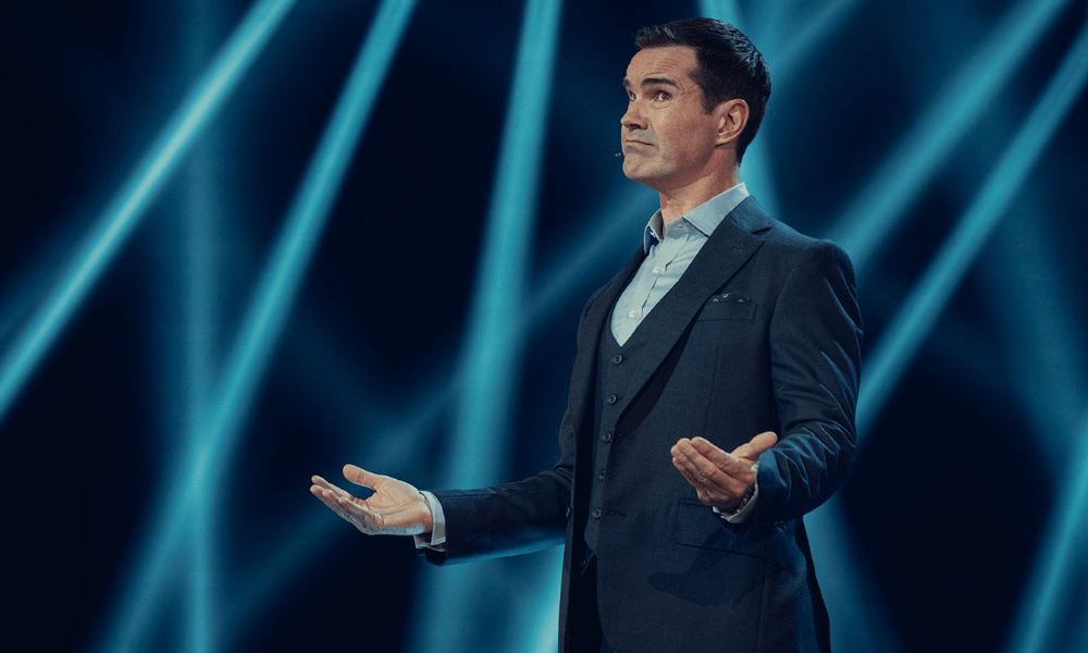 Man in a suit presenting on stage with blue lighting in the background.