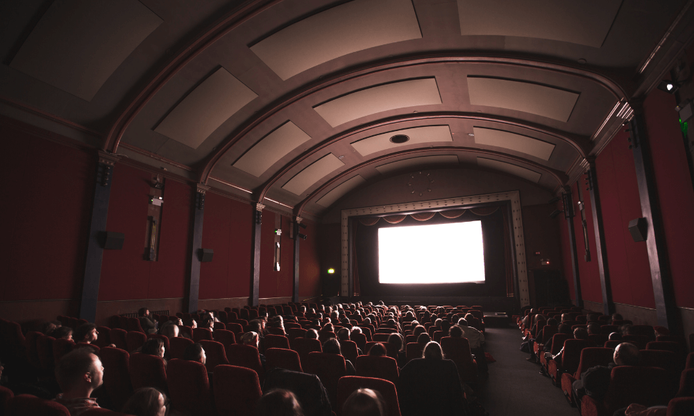 Interior of a dimly lit movie theater with a fully seated audience watching a bright screen.