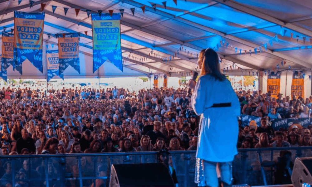 A woman speaks on stage at a crowded festival tent with numerous attendees and blue banners hanging above.