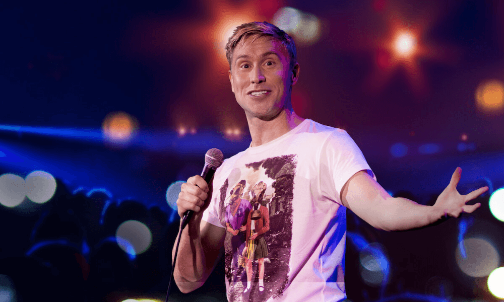 A man with blond hair, wearing a graphic t-shirt, smiling and holding a microphone on stage with a blurred audience and stage lights in the background.