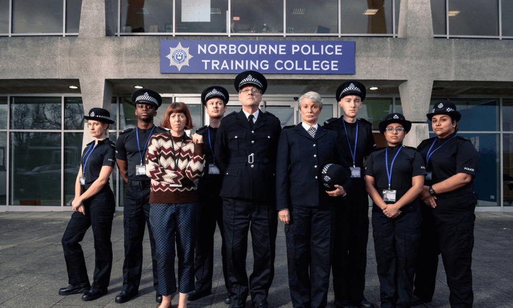A group of nine diverse individuals, some in police uniforms and others in civilian clothes, standing in front of the "norbourne police training college" building.