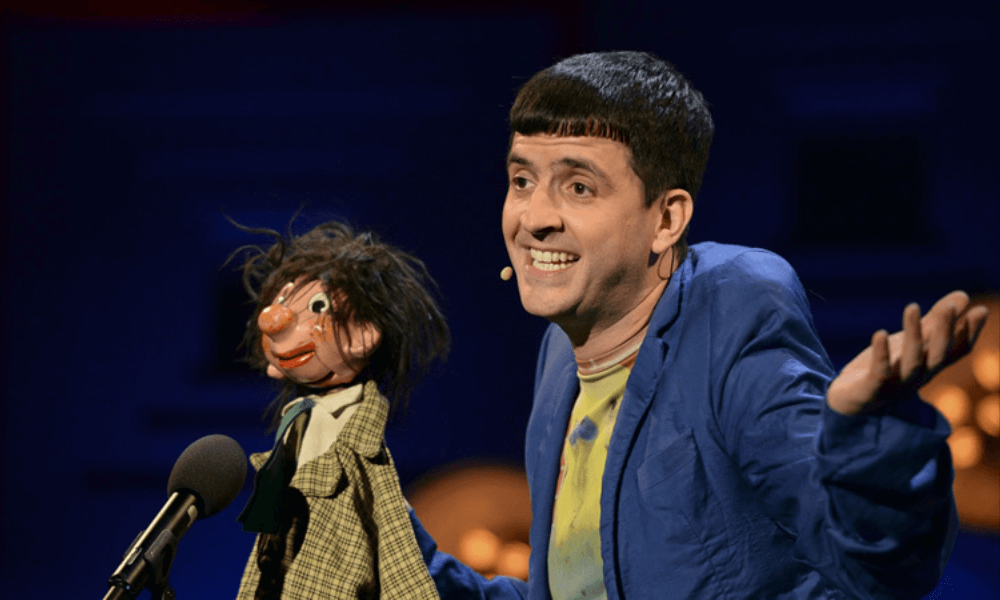 A ventriloquist in a blue jacket performing on stage with his puppet, which wears a plaid jacket.