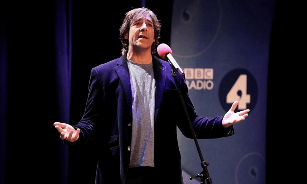A man speaking into a microphone on a stage with a bbc radio 4 backdrop, dressed in a dark blazer and looking expressive.